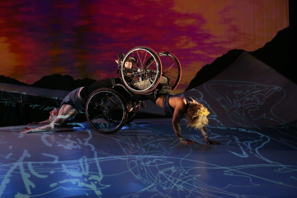 Two dancers, both in wheelchairs; one crawls forward and the other arches her back as she is dragged along the floor. A sunset appears behind them.
