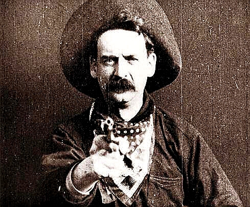 A man dressed as a cowboy wears a wide-brimmed hat and a bandana around his neck. He has a stern expression and a thick mustache and is aiming a revolver directly at the camera, creating a dramatic and confrontational effect. 